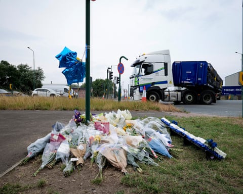 Flowers arranged by roadside with a blue balloon and a blue and white lorry in background.