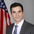 A head and shoulders shot of Gavin Kliger wearing a grey suit, white shirt and blue checked tie, standing next to an American flag against a white wall