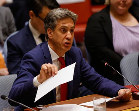 Ed Miliband, the foreign secretary, dressed in a suit and tie talks from behind a table with a microphone on it while waving a piece of white paper.