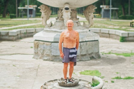A shirtless man playfully stands on a stone water lily flower in the base of a dry fountain.
