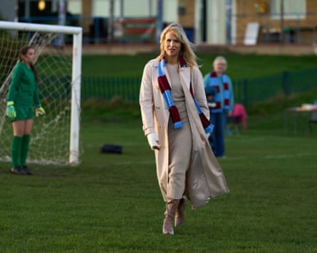 Amanda stands on a football pitch wearing a cream coat and beige outfit, with a blue-and-red team scarf.