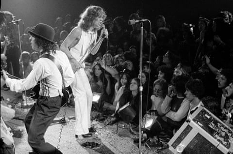 A black and white photo of a long-haired David Johansen singing in front of a crowd in heeled boots, white trousers and a white sleeveless top next to Sylvain Sylvain playing guitar wearing shirt, braces and a bowler hat