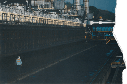 A worker wearing a safety hat and a blue and orange striped shirt walks along machinery at an energy facility