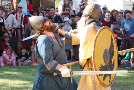 Two bearded men wearing medieval costumes and helmets, fight at close quarters with shields, swords and a dagger. In the background, a a crowd watches.