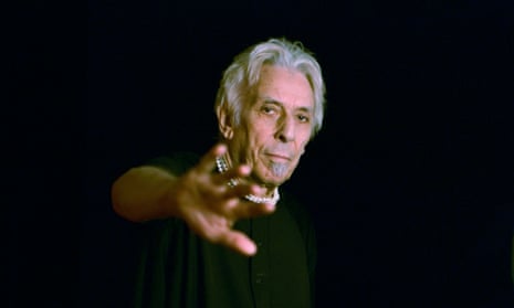 John Cale, white-haired, his arm raised, reaching out to the camera.