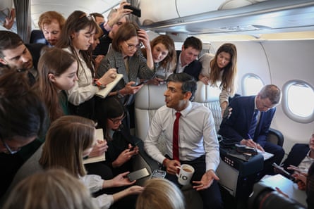 Rishi Sunak sits in a plane seat holding a mug of tea as journalists crowd around him taking notes and holding phones to record what he’s saying