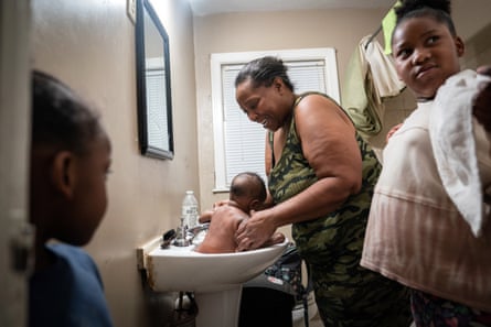 A grandmother bathes a baby in the sink with two other kids in the bathroom with her