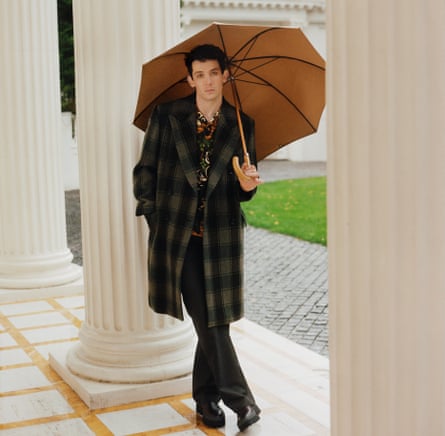 Actor Josh O'Connor standing among pillars at a stately home, holding an umbrella over his head