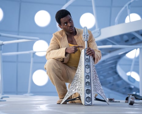 Ncuti Gatwa, in linen suit, rests on one knee and holds and points to at an engrossed silver pyramid
