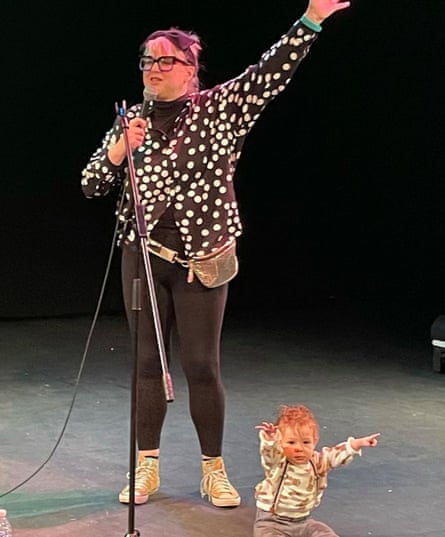 Hatty Ashdown speaking into a mic and raising her hand; a toddler sits on the stage raising their hands towards the audience.