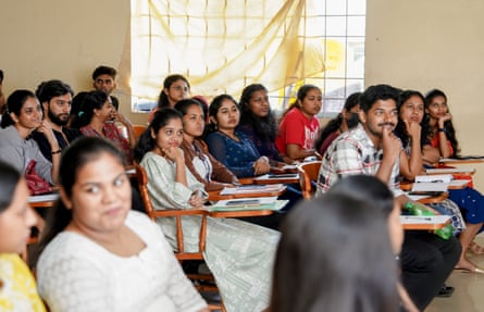 Students sit in rows on chairs with desks attached to one arm.