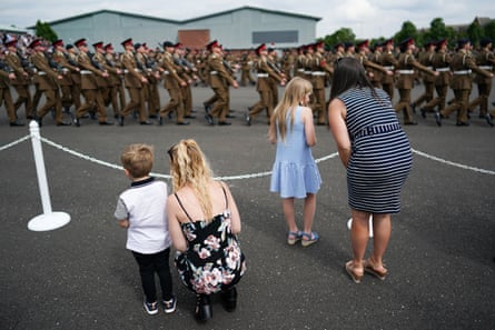 Women with young children stand behind a roped-off area of a parade ground as soldiers in full uniform march past