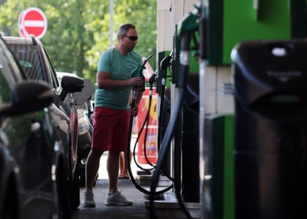 A man lifts a nozzle from a line of pumps to refuel a vehicle at a petrol station in Buckinghamshire. He wears a green T-shirt and red chino shorts, and the petrol pumps are also green.