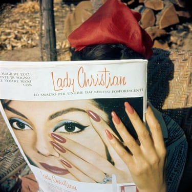 A dark-haired woman in a red hat holds up a magazine she is reading, obscuring her face. The hand holding the magazine has long, painted nails similar to those of the woman pictured on the front of the magazine
