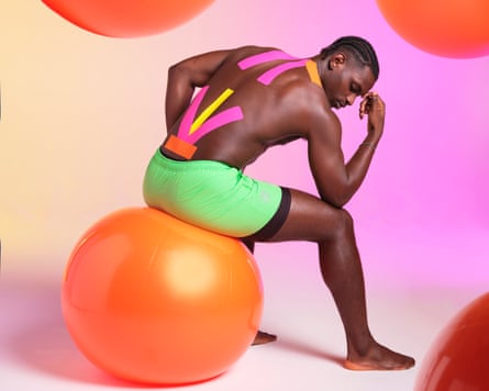 A man seen from the side, wearing green shorts and sitting on an orange exercise ball, with his elbow resting on his knee and his hand against his forehead, with pink, yellow and orange physio tape on his back and neck, with other orange exercise balls around him, against a pink background.