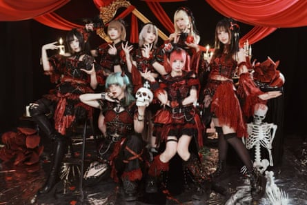 Seven young women in black-and-red goth-style dresses stand in two rows in front of red curtains