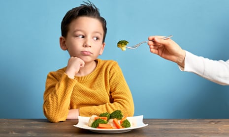 Child sitting at a table, looking grumpy with a plate of vegetables in front of him, and a hand coming into the shot holding a fork with broccoli on it