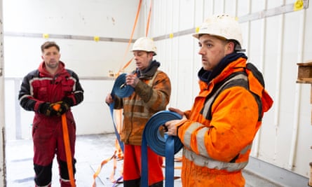 Three men in red or orange waterproofs, two of them in white hard hats, standing in a group