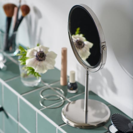 A stainless steel round stand mirror on a green tiled bathroom ledge surrounded by makeup, brushes, jewellery and a single white flower in a clear vase