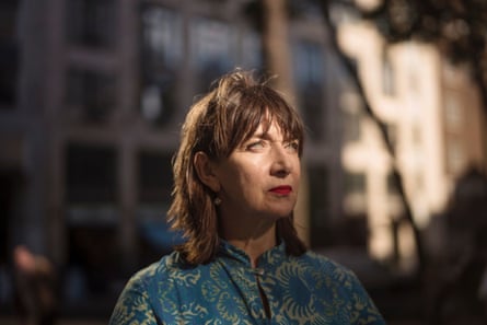 Maureen Beattie in 2018. She wears a blue and gold patterned shirt and red lipstick, and has straight dark hair cut with a fringe. She is standing in part-shadow in front of a building.