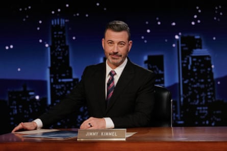 A man sits at a desk on a late-night show set