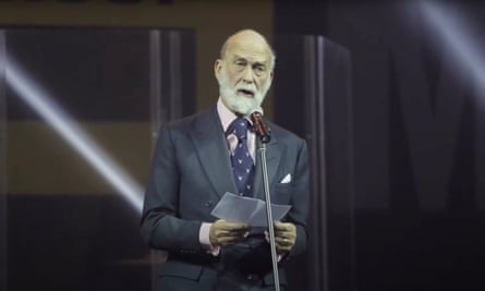 Prince Michael in a business suit, stranding at a microphone, speaking from notes he is holding in his hand