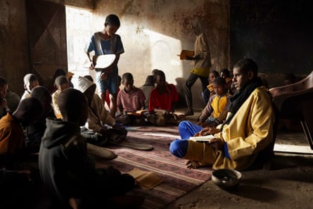 Young African boys are seated on the floor in a circle around a man. Most have books open in front of them, some have wooden boards with writing on them – all with Arabic script