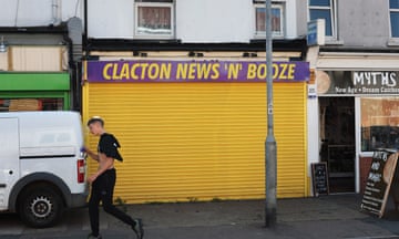 A man with his shirt draped over his shoulder and holding a can of Red Bull walks past the closed Clacton News 'N' Booze shop