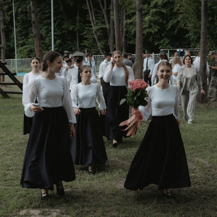 Female cadets walk through a field. They are all wearing identical white blouses and long, flared black skirts. They have their hair tied back and wear makeup; one holds a bunch olf flowers.