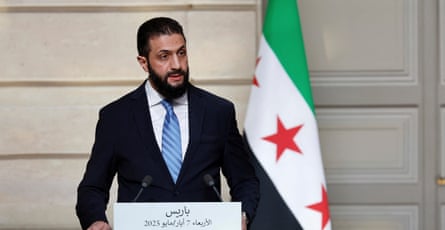 he wears a black suit, white shirt and blue tie, and has a dark beard; he stands in front of a furled Syrian independence flag, which has green, white and black stripes with red stars.