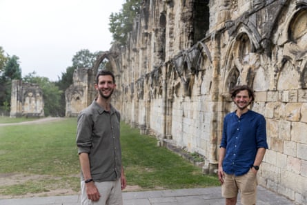 Paul Fawkesley and Ian Drysdale stand against stone walls in Museum Gardens, York; they are young men casually dressed in loose shirts and shorts, smiling widely.