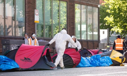 Workers in white overalls and others in hi-vis vests dismantling tents pitched on a street outside a building