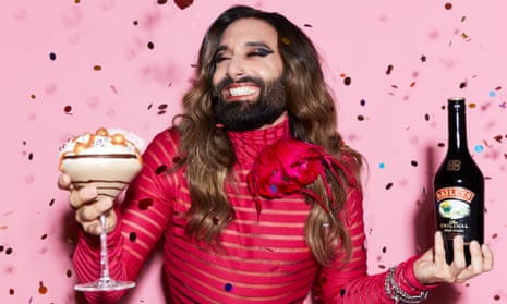Conchita Wurst holding a bottle of Baileys and a large cocktail against pink background