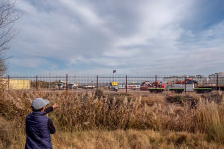 A man standing in an overgrown field pointing to industrial vehicles and machinery parked over a fence