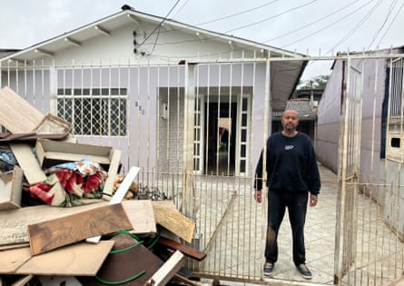 person wearing black sweater and pants stands at gate in front of house with a pile of boards, and items stacked in front of the gate