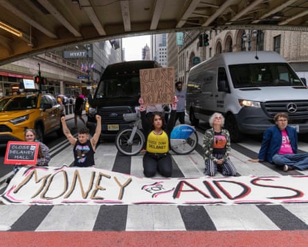 Five people blocking traffic at a crossing on a busy US city road with a banner saying ‘Money for Aids’ and signs and T-shirts saying ‘close Delaney Hall’