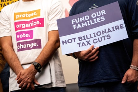 Closeup of people standing at rally, one wearing a white T-shirt saying 'protest. organize. vote. change' and one holding a sign reading 'Fund our families not billionaire tax cuts'