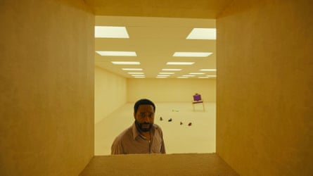 A man stares through an aperture in an queasy yellow room.