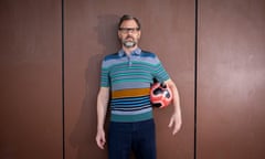 Tim Dowling in a short-sleeved striped polo against a wall holding a football under his arm