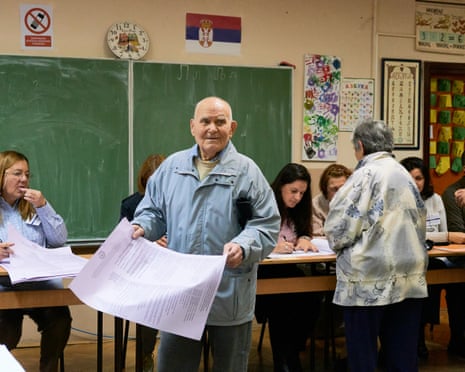 A Serbian voter holds up a large ballot paper at a classroom polling station with a somewhat horrified look on his face.