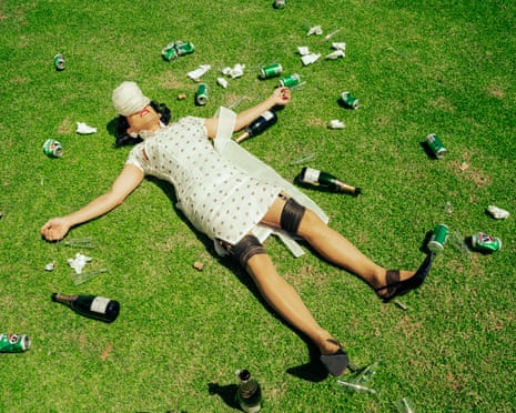 A person in a polka dot dress with a hat on her head lies on grass, surrounded by scattered cans and bottles