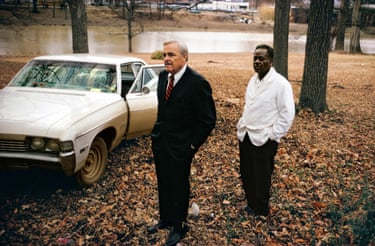 A white man in a dark suit and a person of colour in a white shirt and dark trousers standing in front of a cream coloured cadillac. The ground is full of leaves and there are trees in the background