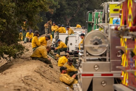 Crowd of men and women dressed in yellow sit on dirt berm next to firetrucks.