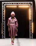 A Black model in a dress with strings and shiny coat with a big collar walks in front of a square steel structure