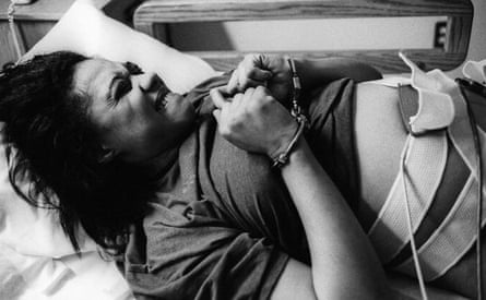 Black and white photograph of a pregnant woman handcuffed in a bed with tubes and straps taped to her stomach, writhing in pain.