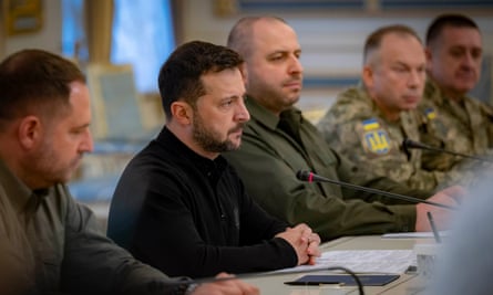 Volodymyr Zelenskyy sits at a long table surrounded by men in khaki and army fatigues