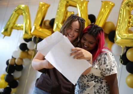Two teenagers open their results together in front of golden balloons that spell out A LEVEL