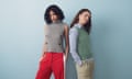 Two models standing together. One has black curly hair and wears a grey sleeveless knit with red tracksuit bottoms. The other has long brown hair and wears a sage green sleeveless knit with a pale blue blouse and chinos.