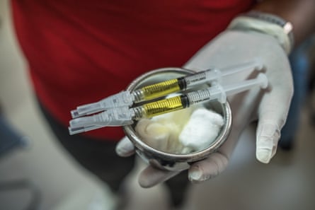 A medical worker holds out two syringes balanced on top of a metal cup containing cotton wool