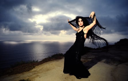 Galas poses in long black gown with a black veil, on a clifftop with dramatic sea behind